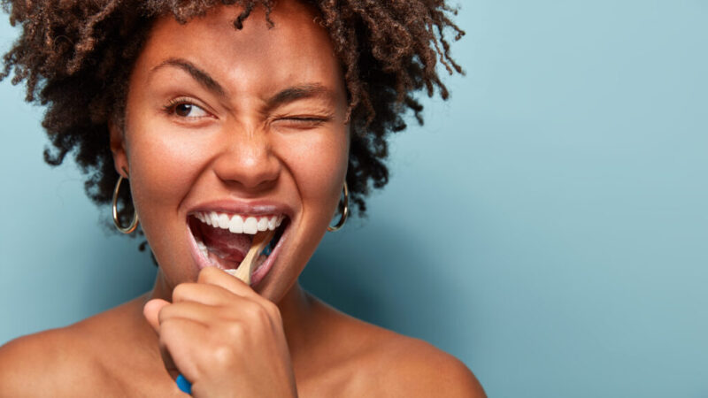 Close up shot of happy joyful funny dark skinned young woman has Afro hair brushes teeth actively with toothbrush, opens mouth broadly, blinks eye, shows bare shoulders, cares of oral hygiene